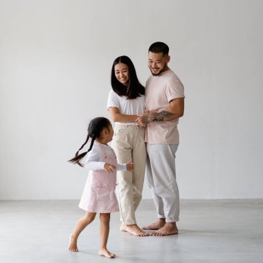 Genç ve gülümseyen çıplak ayaklı ebeveynler el ele tutuşurken küçük kız gri arka planda koşuyor.