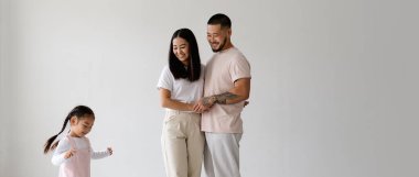 Gülümseyen Asyalı ebeveynlerin el ele tutuşup gri yolda yürümeye başlayan küçük kızın resmi.