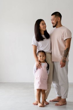 Gülümseyen genç Asyalı ebeveynler, gri üzerinde küçük bir kızın yanında birbirlerine bakıyorlar.