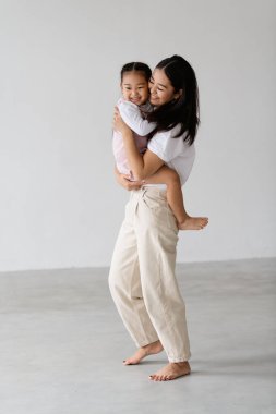 Neşeli ve yalın ayaklı genç Asyalı anne, gri arka planda küçük kızını tutuyor.