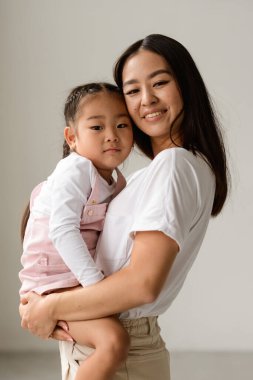 Gülümseyen Asyalı annenin portresi. Küçük kızı kucağında ve gri kameraya bakıyor.