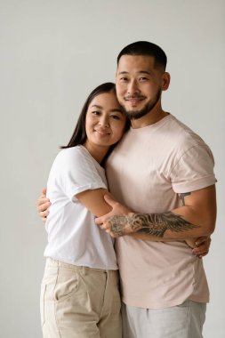 Genç Asyalı tişörtlü bir çiftin portresi. Gri tişörtlerle sarılıp kameraya bakıyorlar.