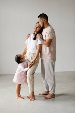 Genç ve yalın ayaklı Asyalı bir aile. Küçük kızı, sarılıyor ve gri arka planda duruyor.