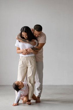 Mutlu genç Asyalı ebeveynler gri arka planda komik küçük kızlarına bakıyor.