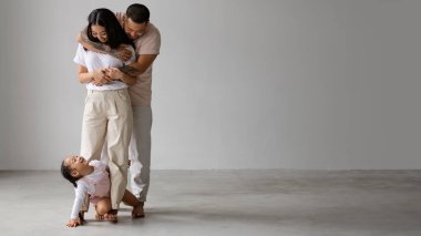 Çıplak ayaklı Asyalı çocuk, gri arka planda sarılan genç ebeveynlerle oynuyor.