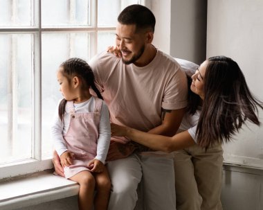 Gülümseyen Asyalı ebeveynler pencerenin kenarında küçük kızlarına sarılıyorlar.