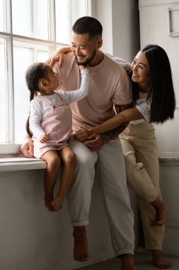 Gülümseyen çıplak ayaklı küçük kız genç Asyalı ebeveynlerin yanında pencere kenarında oturuyor.