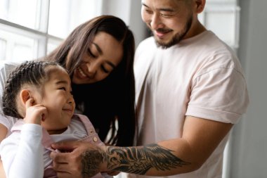 Gülümseyen Asyalı anne-babalar, içeride bulanık pencerenin yanında küçük kızıyla oynuyorlar.