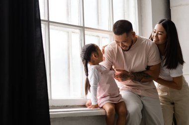 Gülen Asyalı ebeveynler pencerenin kenarındaki küçük çocukla oturuyorlar.