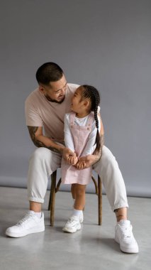 Neşeli Asyalı baba, gri arka planda otururken küçük kızına bakıyor.