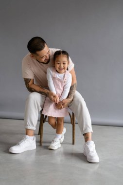 Gülümseyen Asyalı çocuk gri arka planda şık babasının yanında kameraya bakıyor.