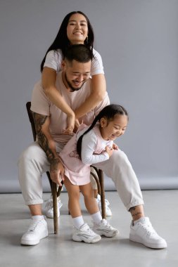 Genç Asyalı kadın kameraya bakıyor ve gri arka planda küçük kızının yanında kocasına sarılıyor.