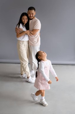 Küçük Asyalı çocuk gülümseyen ebeveynlerin yanında durup gri arka planda sarılıyor.