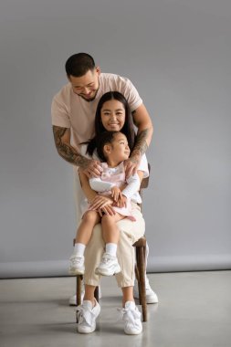 Dövmeli Asyalı adam gri arka planda gülümseyen karısı ve kızına sarılıyor.