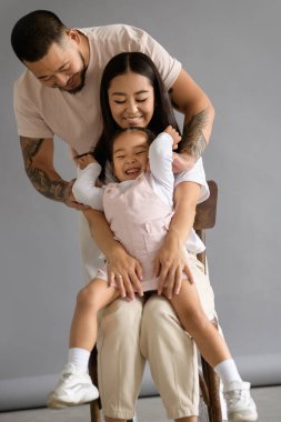 Neşeli Asyalı ebeveynler gri arka planda küçük kızlarıyla oynuyorlar.