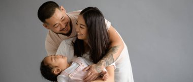 Dövmeli Asyalı adam, kızını kucaklıyor ve gülümseyen eş, gri, afiş manzarasında soyutlanıyor.