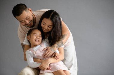 Gülümseyen Asyalı adam gri arka planda kızına ve karısına sarılıyor.