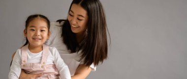 Gülümseyen Asyalı anne, küçük kızına sarılıyor. Gri, afiş manzarasında izole edilmiş.