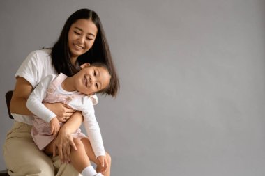 Gülümseyen Asyalı anne, gri arka planda izole edilmiş küçük kızına sarılıyor.