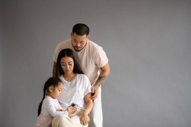 Genç Asyalı ebeveynler gri arka planda izole edilmiş akıllı telefonlu küçük kızlarına bakıyorlar.