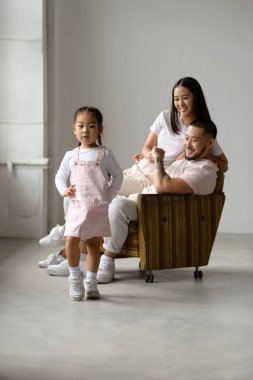 Gülümseyen ebeveynlerin yanında poz veren Asyalı küçük kız gri arka planda koltukta oturuyor.