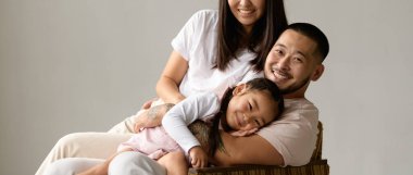 Neşeli Asyalı genç adam karısına sarılıyor ve küçük kızı da gri stüdyoda izole edilmiş.