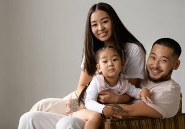 Gülümseyen Koreli ebeveynler gri arka planda izole edilmiş küçük kızın yakınındaki kameraya bakıyorlar.