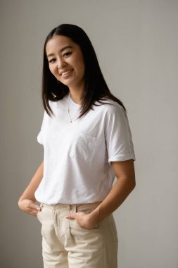 Gri arka planda izole edilmiş kameraya bakan sıradan giyinmiş pozitif Asyalı kadın portresi