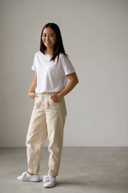 Gri arka planda dikilirken kameraya bakan günlük giysili genç Asyalı kadın.