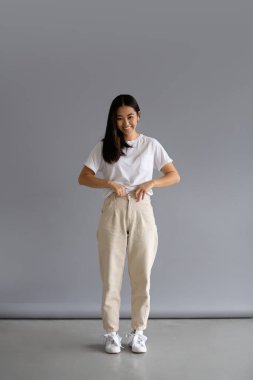 Neşeli genç Asyalı kadın pantolonunu düzeltiyor ve gri arka plandaki kameraya bakıyor.