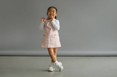Sundress giymiş neşeli Asyalı çocuk kameraya bakıyor ve gri arka planda duruyor.