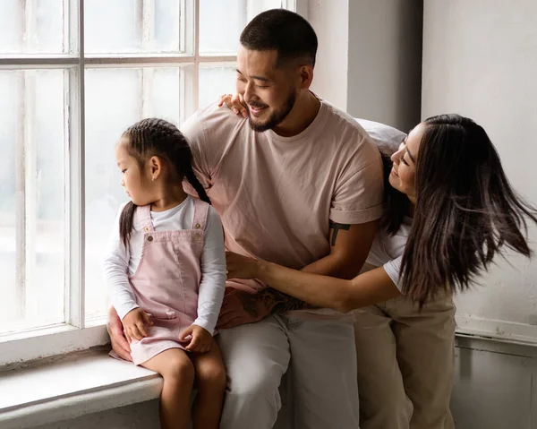 Gülümseyen Asyalı ebeveynler pencerenin kenarında küçük kızlarına sarılıyorlar.