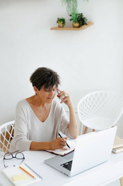 Evde akıllı telefondan konuşan ve not defterine yazan olgun bir serbest yazar.