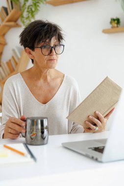 Evde dizüstü bilgisayarın yanında gözlüklü, elinde kitap ve kahve tutan olgun bir kadın.