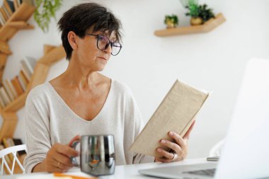 Evde laptopun yanında kitap okuyan olgun bir kadın.