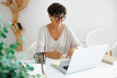 Evde çalışan, kulaklıklı, gülümseyen, olgun bir serbest çalışan.