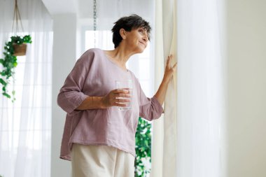 Sabahları pencerenin yanında su bardağı tutan olgun bir kadın.