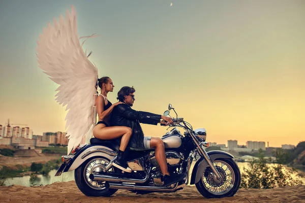 Jovem casal bonito elegantemente vestido em uma motocicleta ao pôr do sol, em uma mulher asas de anjo, anjo da guarda, condução perigosa — Fotografia de Stock