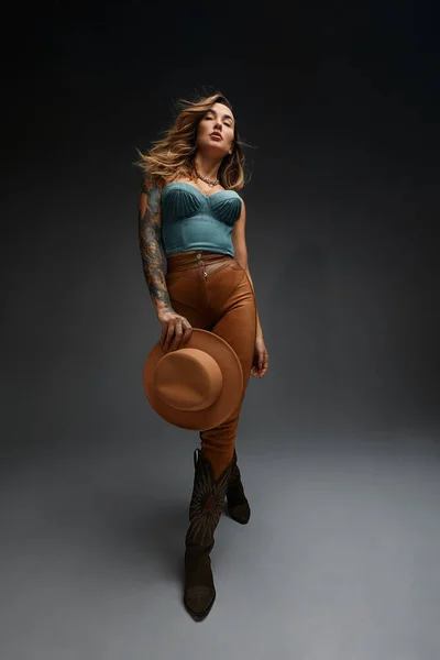 Retrato de moda de chica morena joven en top occidental azul y jeans con sombrero de vaquero y botas en frente de fondo de estudio de copia gris espacio. Retrato de moda y estilo de vida. —  Fotos de Stock