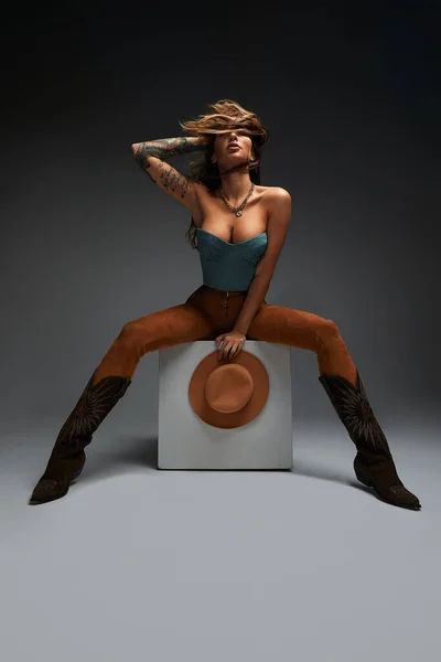 Mode porträtt av ung brunett flicka i blå västra toppen och jeans med cowboy hatt och stövlar framför grå kopia utrymme studio bakgrund. Mode porträtt och livsstil. — Stockfoto