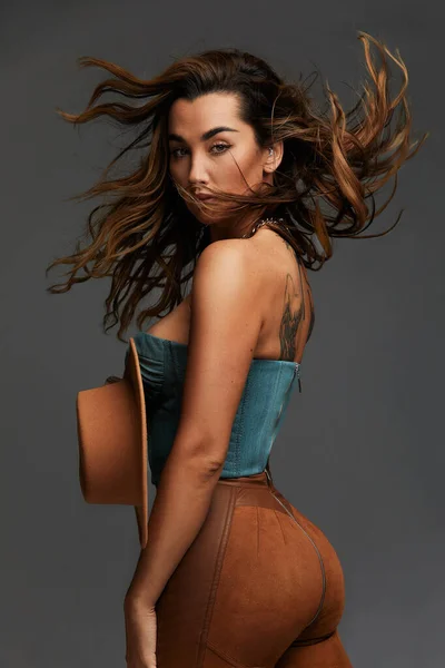 Retrato de moda de chica morena joven en top occidental azul y jeans con sombrero de vaquero y botas en frente de fondo de estudio de copia gris espacio. Retrato de moda y estilo de vida. —  Fotos de Stock