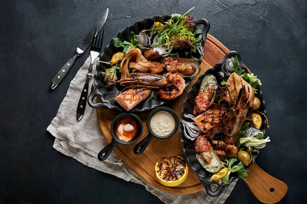 Set de platos de mariscos asados mixtos. Contienen camarones grandes a la parrilla, calamares y Barracuda Pimiento de ajo de pescado con salsa de chile picante y papas, sobre tabla de madera y fondo negro. — Foto de Stock