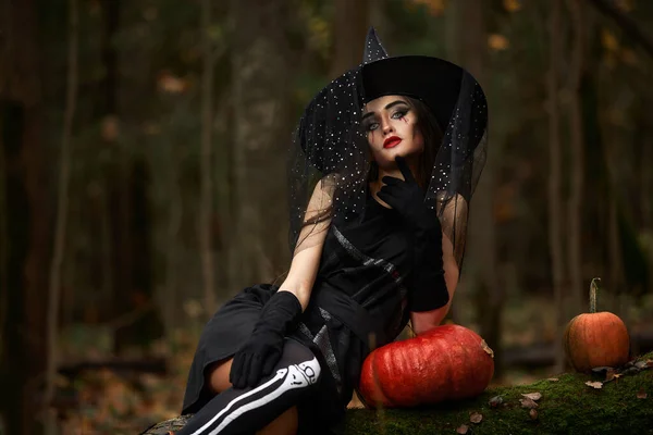 Mujer joven en vestido negro con sombrero de bruja y calabaza naranja colocada en el bosque, concepto de Halloween. Tema Horror. — Foto de Stock