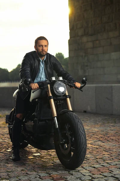Snygg fashionabla cyklist klädd i en svart läderjacka sitter på sin motorcykel, tittar på kameran. — Stockfoto
