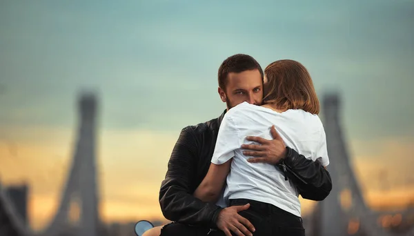 Jong sexy vrouw knuffelen schattig man in stijlvolle zwart lederen jas, zittend op sport motorfiets op de brug in de stad op zonsondergang en zoenen — Stockfoto