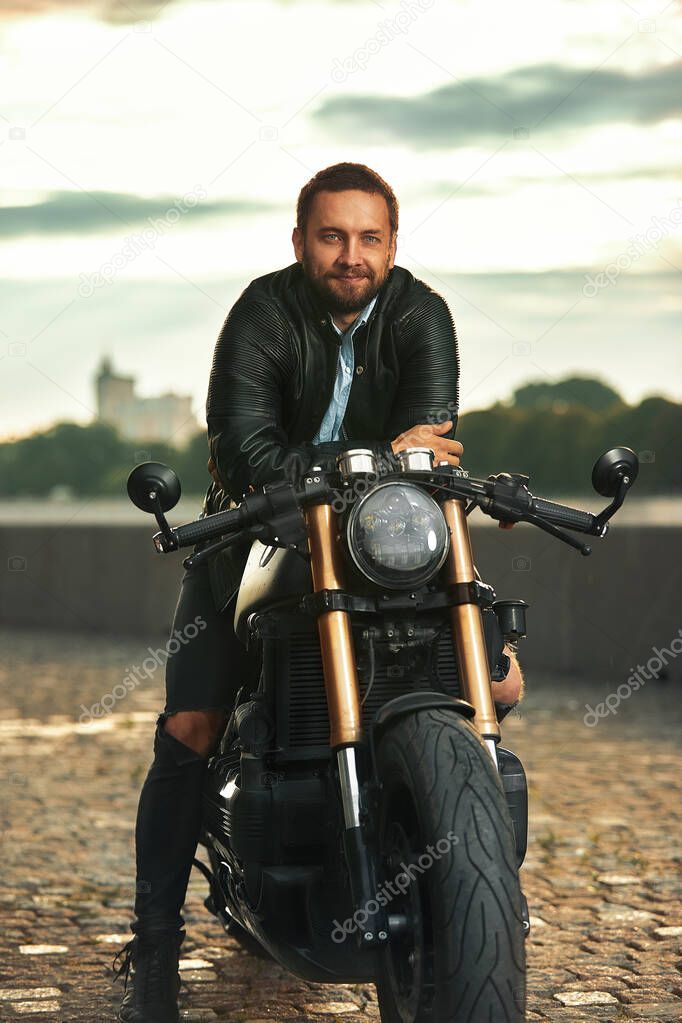 Stylish fashionable biker dressed in a black leather jacket sitting on his motorcycle, looking at camera.