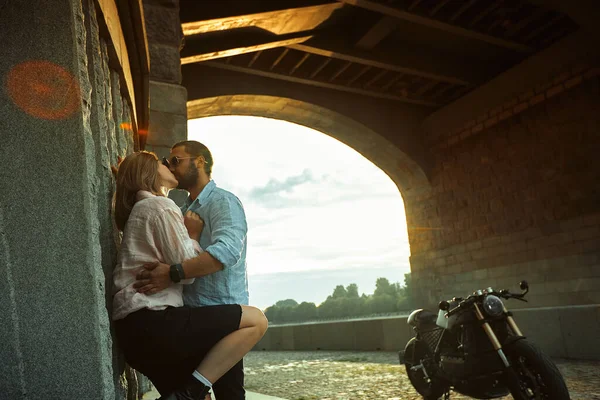 Liefde en romantisch concept. Paar verliefd op zonsondergang staan bij de muur kus en knuffel onder de brug naast de motor. Man met baard knuffelende vrouwen, tederheid — Stockfoto