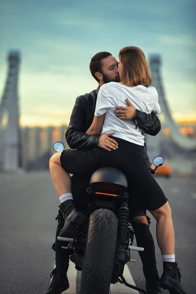Joven mujer sexy abrazando hombre lindo en chaqueta de cuero negro con estilo, sentado en motocicleta deportiva en el puente de la ciudad en la puesta del sol y besar — Foto de Stock