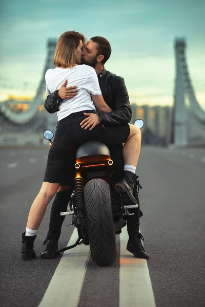 Jong sexy vrouw knuffelen schattig man in stijlvolle zwart lederen jas, zittend op sport motorfiets op de brug in de stad op zonsondergang en zoenen — Stockfoto