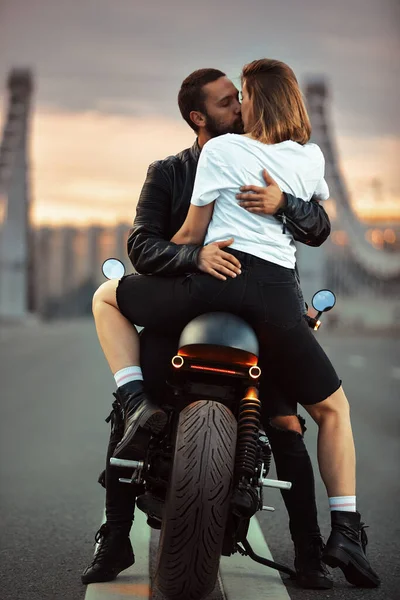 Jovem mulher sexy abraçando o homem bonito na jaqueta de couro preto elegante, sentado na motocicleta esportiva na ponte da cidade no pôr do sol e beijando — Fotografia de Stock